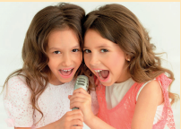 Imagem: Fotografia. Duas meninas sorriem enquanto cantam juntas segurando um microfone. Fim da imagem.