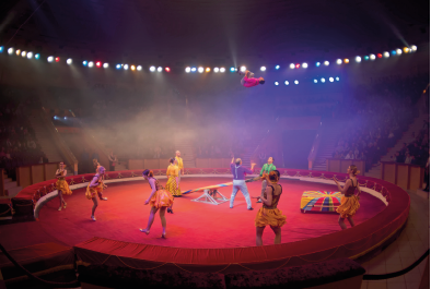 Imagem: Fotografia. Interior de uma tenda de circo. É escura com pontos de iluminação circular no teto e um grupo com cerca de dez pessoas com roupas coloridas ao centro do picadeiro. O chão é vermelho, ao centro há uma gangorra e uma pessoa faz cambalhota no ar.  Fim da imagem.