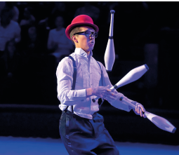 Imagem: Fotografia. Um rapaz de calça com suspensório, camisa, chapéu vermelho e óculos faz malabarismo com três pinos.  Fim da imagem.