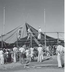 Imagem: Fotografia em preto e branco. Em um espaço aberto, um grupo de pessoas monta um circo com lona e uma estrutura circular montada.  Fim da imagem.