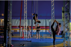 Imagem: Fotografia. Em um amplo espaço fechado com muitas colunas verticais de ferro está um grupo de quatro meninas e uma mulher. Em primeiro plano, uma menina faz alongamento com uma das pernas atrás da cabeça. Ao fundo, uma delas está de ponta cabeça em uma trave. Ao lado, as demais observam.  Fim da imagem.