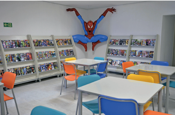 Imagem: Fotografia. Uma sala que apresenta mesas e cadeiras coloridas. Ao fundo, prateleiras com muitos gibis e a figura do personagem Homem-Aranha em tamanho grande representado na quina das paredes. Fim da imagem.