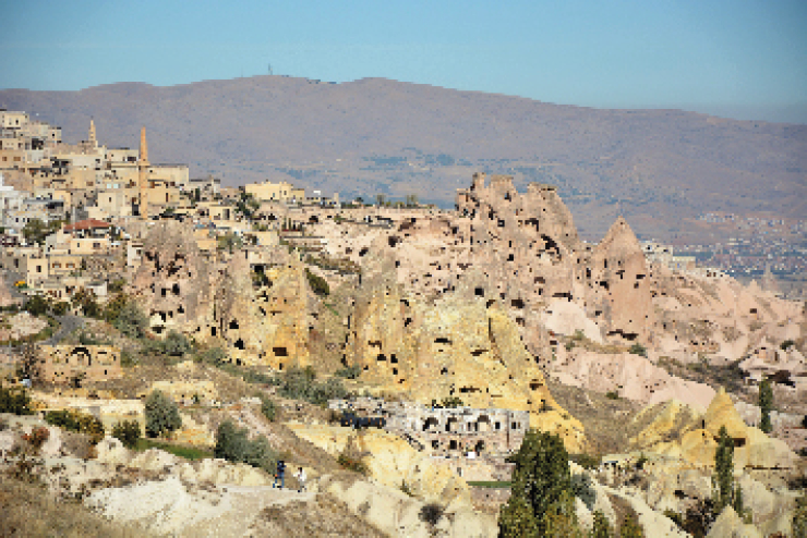 Imagem: Fotografia. Vista panorâmica de um vale com rochas escavadas com aberturas de portas e janelas e algumas casas em meio à pouca vegetação. No horizonte, montanha e o céu.  Fim da imagem.