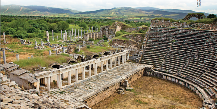 Imagem: Fotografia. Vista panorâmica de um local aberto com ampla construção em ruínas. Apresenta uma alta arquibancada semicircular, um palco comprido com colunas dispostas na parte de trás. Ao redor, restos de colunas e muros em uma área gramada. No horizonte, montanhas.  Fim da imagem.