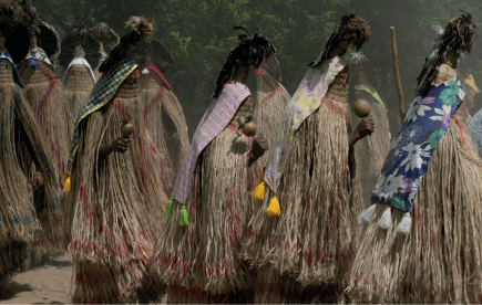 Imagem: Fotografia. Destaque de uma região aberta com chão de areia e fundo com árvores e por onde caminham personagens com corpo todo coberto com longas franjas bege com trechos tingidos de rosa e azul e tecidos estampados na parte de trás do corpo e presos em um adereço de penas na cabeça. As mãos estão de fora e seguram maracás.  Fim da imagem.