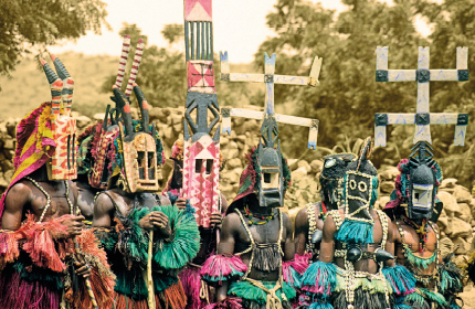 Imagem: Fotografia. Em um lugar aberto com árvores, destaque com seis homens negros que vestem figurinos coloridos com franjas nos braços, tórax e cintura. Eles usam adereços no pescoço e máscaras em formato geométrico e que cobrem todo o rosto e com estruturas na parte superior, como um chifre e símbolos.  Fim da imagem.