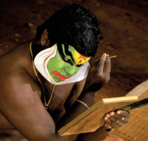 Imagem: Fotografia. Um homem sem camisa está sentado e usa uma máscara verde com pintura em cor preta, amarela, vermelha e com rebarbas brancas na lateral. Ele usa um colar, pulseira e lê um livro aberto em sua frente.  Fim da imagem.