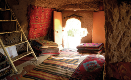 Imagem: Fotografia. Interior de uma caverna onde há uma escada de madeira, tapete estampado, assentos com almofadas e a porta com entrada da luz solar.  Fim da imagem.