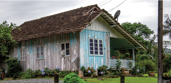 Imagem: Fotografia. Fachada de uma casa de madeira com janelas de vidro, área com plantas e quintal gramado. O teto é de telhas e tem formato de “V” invertido.  Fim da imagem.