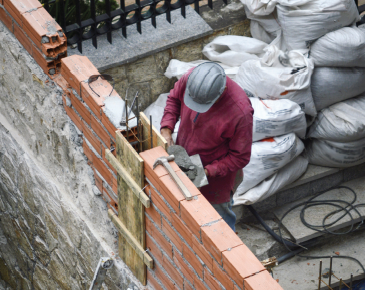 Imagem: Fotografia. Um homem de capacete está diante de um muro de tijolos em construção. Atrás dele, estão muitos sacos de cimento.  Fim da imagem.