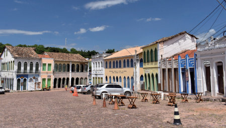 Imagem: Fotografia. Destaque de uma área aberta com chão de pedras e cercada por casas e estabelecimentos com fachadas assobradadas com muitas janelas e portas de acesso voltado diretamente à rua. No local, há carros estacionados e mesas de madeira dispostas na via.  Fim da imagem.