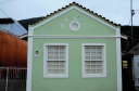 Imagem: Fotografia. Em uma área cercada de outras construções, fachada de uma casa de alvenaria com janelas de vidro. Fim da imagem.