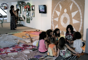 Imagem: Fotografia. Em uma sala, um pequeno grupo de crianças está sentado em roda junto de uma mulher adulta. Eles estão sobre tecidos estendidos no chão e na parede há um desenho circular em mosaico. Ao fundo, outras pessoas de pé numa área onde há objetos expostos na parede.  Fim da imagem.