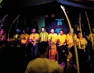 Imagem: Fotografia. Um homem está em destaque à frente de um grupo de instrumentistas uniformizados, dentre eles muitos estão com berimbaus.  Fim da imagem.