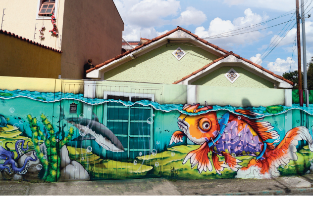 Imagem: Fotografia. Destaque do muro e portão de uma casa que estão com uma pintura do fundo mar, com um grande peixe laranja e roxo, tubarão, polvo, algas e pedras. A casa é térrea e ao redor há outras residências e postes com fiação. Fim da imagem.