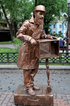 Imagem: Fotografia. Em uma rua, um homem parado de pé sobre uma mala deitada no chão e apoiado em uma caixa sobre uma base vertical. Ele usa longo casaco, chapéu e possui barba comprida. Sua roupa, pele e objetos tem aspecto de bronze. Fim da imagem.