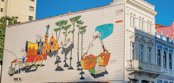 Imagem: Fotografia. Destaque de uma pintura no muro lateral de um edifício. Na pintura, há uma mulher com roupa longa e larga e tecido amarrado na cabeça. Ela caminha segurando dois cestos cheios de frutas próxima de um lago e de árvores. Ao fundo, uma igreja e uma carroça onde um homem está sentado e aguarda o cavalo beber água. Ao redor desse edifício, há outros prédios e árvore.  Fim da imagem.