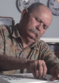 Imagem: Fotografia. Retrato de um homem calvo com bigode curto grisalho e que usa camisa. Ele está com a cabeça inclinada para o lado esquerdo e olha para baixo. Fim da imagem.