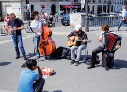 Imagem: Fotografia. Em uma rua, durante o dia, um grupo de quatro músicos com violão, sanfona, contrabaixo e flauta se apresenta diante de um pequeno cesto disposto à frente deles. Um menino sentado na rua os observa. Ao fundo, pessoas transitam. Fim da imagem.