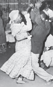 Imagem: Fotografia em preto e branco. Um homem e uma mulher negros, ele paletó e ela de vestido longo e turbante, dançam com as barrigas unidas e os braços para o alto.  Fim da imagem.