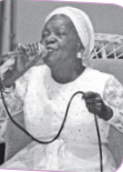 Imagem: Fotografia em preto e branco. Uma senhora negra com vestido e turbante claros está sentada e canta segurando o microfone. Fim da imagem.