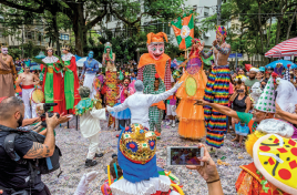 Imagem: Fotografia. Em um espaço aberto, duas pessoas dançam ao centro próximo a um grupo de bonecos gigantes com fantasias coloridas. Fim da imagem.