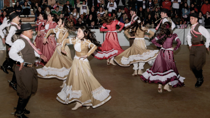 Imagem: Fotografia. Destaque de dançarinos que se apresentam em um amplo espaço com plateia ao fundo. Eles dançam em par. As mulheres usam longo vestidos rodados e sapatos e os homens, que estão à sua frente enfileirados, usam chapéu, colete, camisa de manga longa, calça e botas de cano longo.  Fim da imagem.