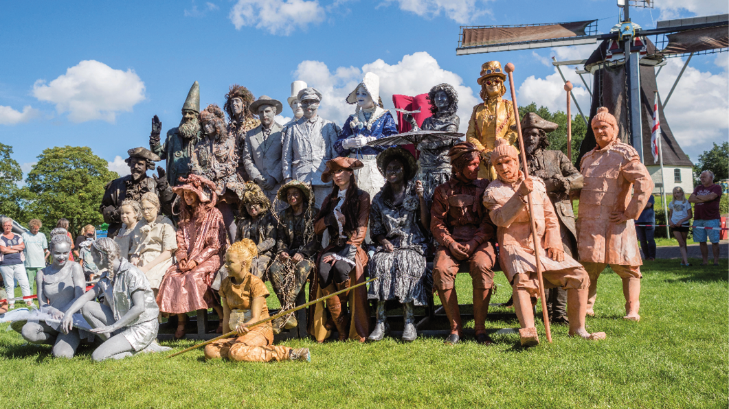 Imagem: Fotografia. Em um espaço aberto gramado, durante o dia, um grupo de pessoas com cabelo e rosto pintados e com figurinos diversos, como duende, piratas, marinheiro, bonecos e espantalho, posam para uma foto. Fim da imagem.