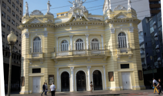 Imagem: Fotografia. Fachada de um edifício de dois andares e que apresenta entrada com três arcos no piso térreo. No piso superior, há cinco janelas arqueadas com vidros e pequenas sacadas.  Na parte superior, há pequenas esculturas. Ao redor, há prédios e pedestres.  Fim da imagem.