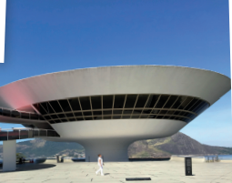 Imagem: Fotografia. Fachada de um prédio que apresenta arquitetura com linhas curvas e superfície com janelas amplas envidraçadas que circundam toda estrutura. Do lado esquerdo, há uma passarela que parte do edifício e se estrutura sobre pilares. Ao redor, um amplo espaço aberto onde caminham algumas pessoas. Ao fundo, morros.  Fim da imagem.