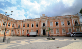 Imagem: Fotografia. Fachada de um amplo prédio com estrutura retangular e com dois pisos com muitas janelas envidraçadas e colunas na entrada. Ao redor, um amplo espaço aberto.  Fim da imagem.