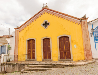 Imagem: Fotografia. Fachada de uma construção térrea que apresenta três amplas portas de madeira e um vitral em formato de cruz na parte superior central.  A rua é composta por paralelepípedos.  Fim da imagem.