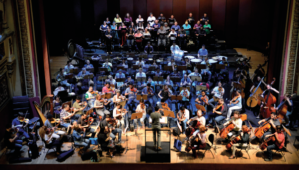 Imagem: Fotografia. Instrumentistas de uma orquestra tocam em um palco. Eles estão unidos em diferentes grupos que compõem um todo e estão diante de uma pessoa que está de pé em uma estrutura mais alta e à frente de partituras.  Fim da imagem.