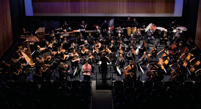 Imagem: Fotografia. Vista panorâmica de um palco em um espaço fechado onde uma orquestra se apresenta. Há instrumentos de corda, percussão e metais. Todos usam roupas escuras e à frente do grupo está um homem de pé em uma estrutura destacada e diante de uma partitura. Na plateia, que está escura, muitas pessoas observam.  Fim da imagem.