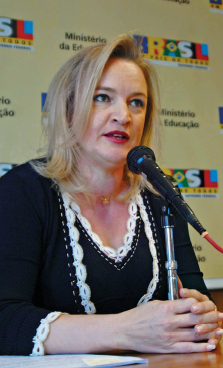 Imagem: Fotografia. Retrato de uma mulher de pele clara, cabelo liso loiro, maquiada e que está sentada diante de um microfone. Ao fundo, um painel onde está escrito “Ministério da Educação” e “Brasil”. Fim da imagem.