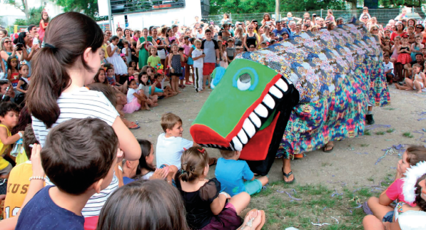 Imagem: Fotografia. Em uma área aberta, muitas crianças estão reunidas em roda ao redor de pessoas enfileiradas que vestem conjuntamente o figurino de um animal com cabeça grande com dentes afiados e corpo comprido com capa florida. Na apresentação, o animal está com a boca próxima da cabeça de uma criança sentada no chão.  Fim da imagem.