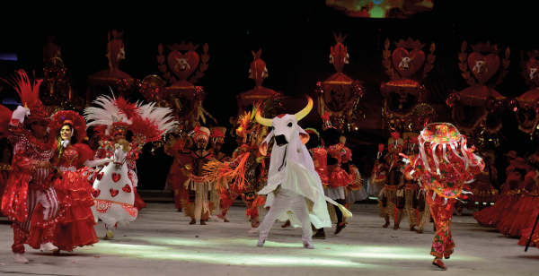 Imagem: Fotografia. Em um palco iluminado um grupo de dança se apresenta. Ao centro, está uma pessoa vestida de branco e com o figurino de um boi branco com chifres longos e afiados. Ao redor, pessoas vestidas com figurino vermelho com adornos brilhantes, com fitas, babados e penas.  Fim da imagem.