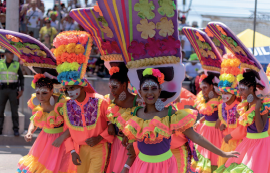 Imagem: Fotografia. Destaque de um grupo de pessoas que desfilam com figurinos de cores vivas em roxo, amarelo, rosa e verde. As mulheres usam babados, saias rodadas com adereço longo na cabeça e o os homens usam adorno volumoso colorido na cabeça, colete e calça. Fim da imagem.