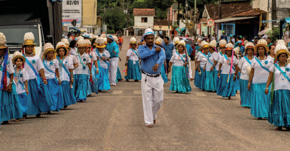 Imagem: Fotografia. Na rua, durante o dia, destaque de um grupo que desfila com roupa azul e branco. Ao centro, está um homem de chapéu, camisa e calça. Ao redor, formando duas fileiras diagonais que se encontram ao fundo, estão mulheres que usam camiseta, saia rodada e chapéu adornado na cabeça. Ao fundo, casas.  Fim da imagem.