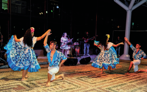 Imagem: Fotografia. Em um palco, há o destaque de dois casais dançando. Eles vestem azul e branco. As mulheres usam saia rodada, camisa e estão de pé com uma das mãos dadas aos homens, que estão ajoelhados e usam camisa e bermuda. Ao fundo, a banda.  Fim da imagem.