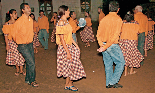 Imagem: Fotografia. Em uma área aberta, durante a noite, um grupo de pessoas, homens e mulheres, dança formando uma roda. Eles usam camisa e calça e estão com as mãos para trás e elas usam camiseta presa no ombro, estão com as mãos na saia longa e usam sandália. Fim da imagem.