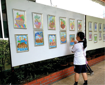 Imagem: Fotografia.  No pátio, uma menina uniformizada observa uma exposição de quadros coloridos dispostos em um extenso painel.  Fim da imagem.