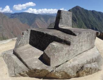 Imagem: Fotografia. Destaque de uma grande composição de pedra robusta iluminada pelo sol e que apresenta uma parte verticalizada que forma um pouco de sombra. Ao fundo, uma extensa cadeia montanhosa e o céu.  Fim da imagem.