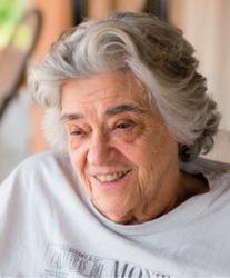 Imagem: Fotografia. Retrato de uma senhora sorridente de pele clara com muitas rugas e cabelo liso grisalho e curto. Ela olha para o lado e usa camiseta.  Fim da imagem.