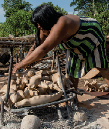 Imagem: Fotografia. Em um local aberto, uma mulher de vestido listrado manipula pequenas esculturas de cerâmica em formato de animais e que estão sobrepostas sobre um forno artesanal feito com base de ferro. Ao fundo, outras peças estão dispostas no chão e há muitas árvores. Fim da imagem.