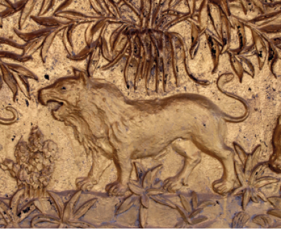 Imagem: Fotografia. Em tons de bege e marrom, destaque de uma parede na qual está entalhada a figura de um leão de pé com a boca aberta ao redor de plantas. Fim da imagem.