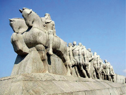 Imagem: Fotografia. Em um local aberto, uma estátua robusta sobre uma ampla base retangular onde estão representados dois homens a cavalo e atrás deles duas compridas fileiras de homens que marcham seminus e descalços.  Fim da imagem.