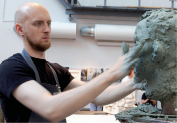 Imagem: Fotografia. Um homem que usa jaleco está com expressão séria enquanto esculpe um busto de argila. Fim da imagem.