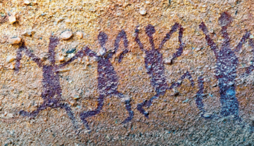 Imagem: Fotografia. Destaque de uma parede rochosa onde estão pintadas silhuetas de quatro figuras humanas lado a lado com a mãos levantadas. Fim da imagem.