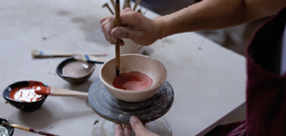 Imagem: Fotografia. Destaque das mãos de uma pessoa que usa jaleco e pinta uma pequena peça de cerâmica com pincel sobre uma bancada. Na mesa, há frascos de tinta e pincéis. Fim da imagem.